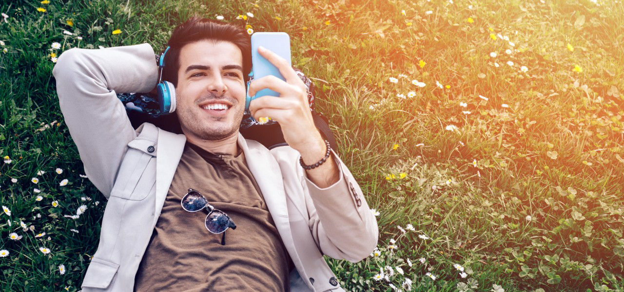 Mann liegt im Gras und hört mit Kopfhörern Musik von seinem Mobiltelefon