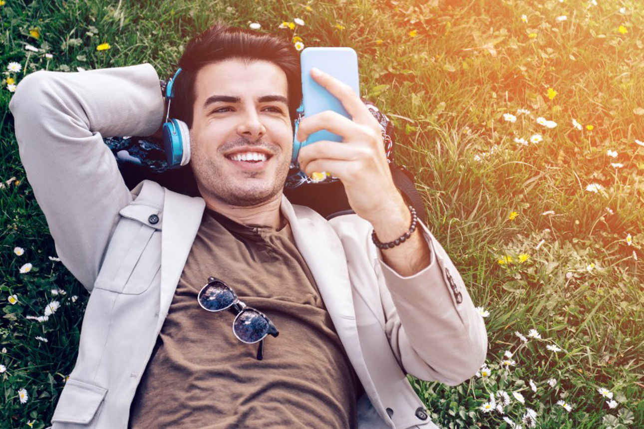 Mann liegt im Gras und hört mit Kopfhörern Musik von seinem Mobiltelefon