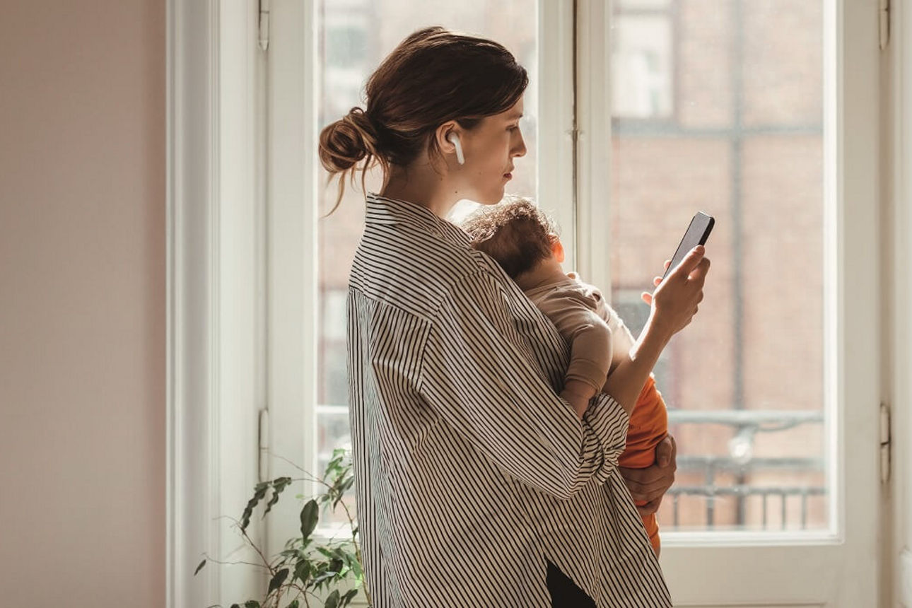 Eine Mutter steht mit ihrem Baby auf dem Arm am Fenster und erledigt ihre Finanzen über die App. 