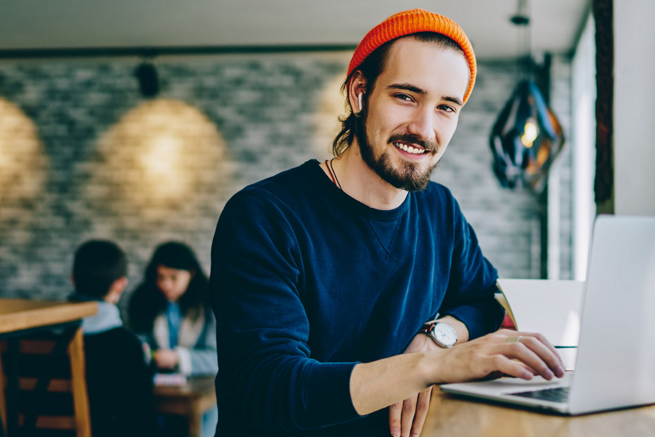 Junger Mann sitzt vor einem Laptop in einem Cafe und lächelt