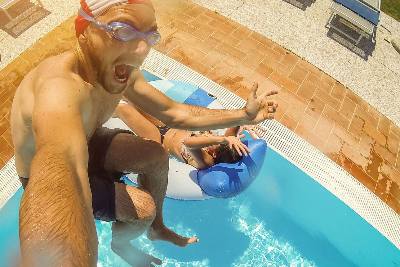Mann in Badesachen macht ein Selfie während er in einen Pool springt.