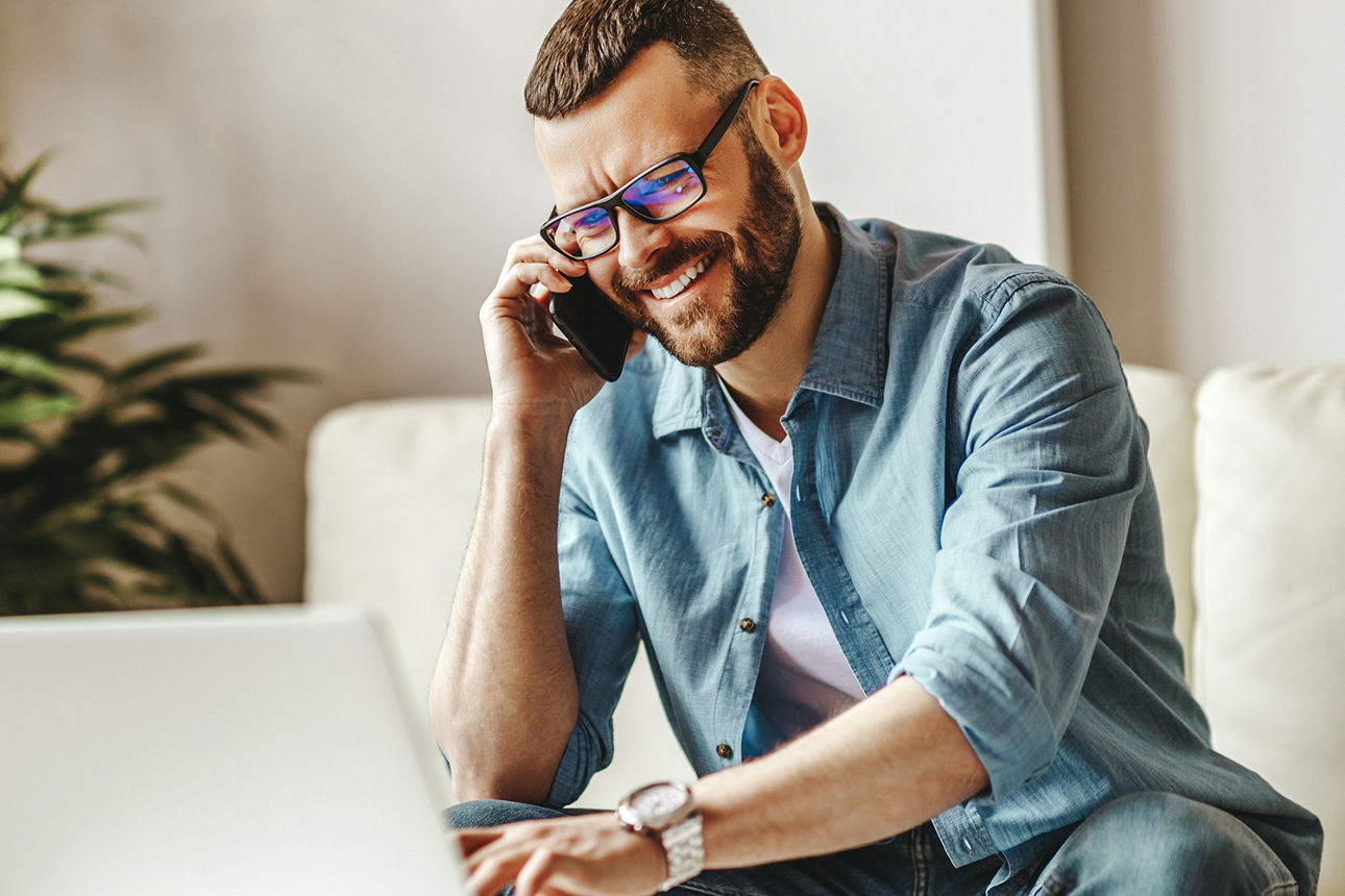 Junger Mann sitzt mit einem Laptop auf dem Sofa unt telefoniert mit einem Smartphone