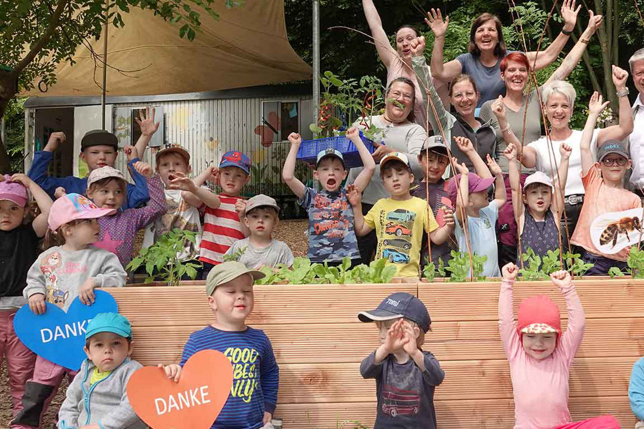 Eine Gruppe Kinder und Erwachsener posiert fröhlich hinter und vor einem Holzkasten mit Pflanzen. Einige halten Schilder mit 