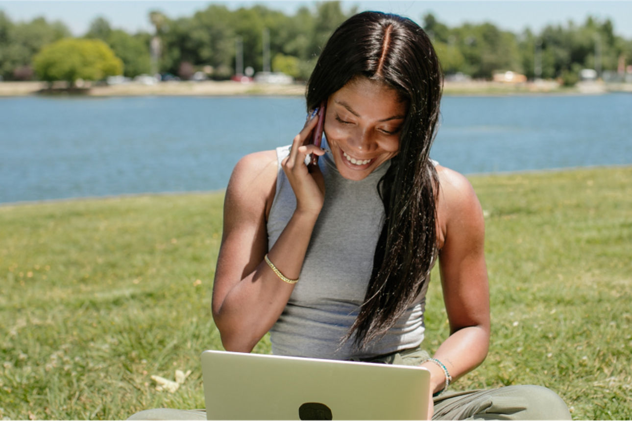 Frau sitzt im Park, hat ein Laptop auf dem Schoss und telefoniert lächelnd