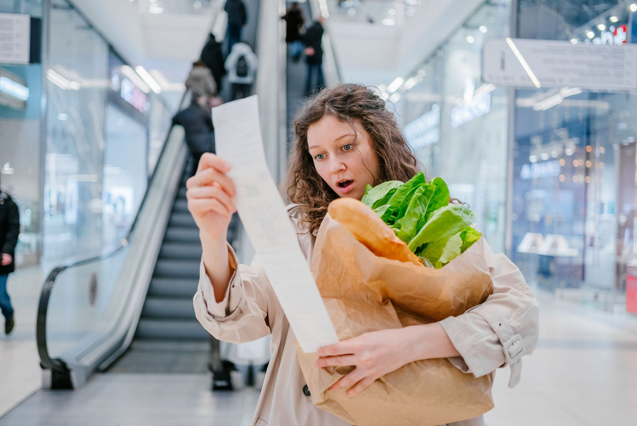 Eine Frau mit einer Einkaufstüte in der Hand, schaut erschrocken auf den Kassenbon