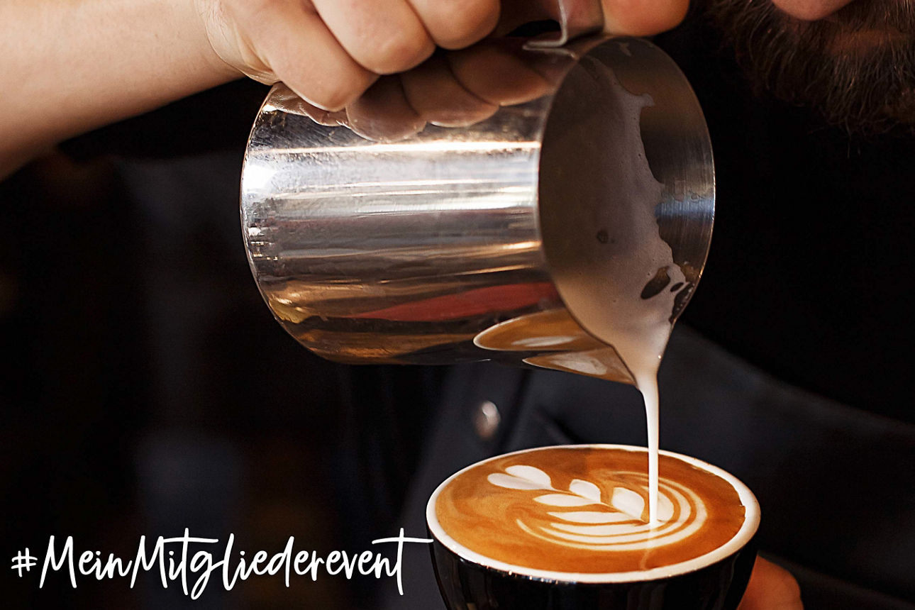 Eine Person gießt Milchschaum aus einem Metallkännchen in eine Tasse Kaffee und formt ein Herzmuster auf dem Cappuccino