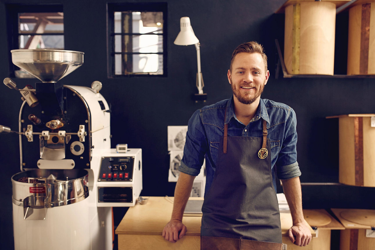 Mann mit Schürze lehnt an einen Tresen. Neben ihm steht eine Kaffeeröstmaschine.