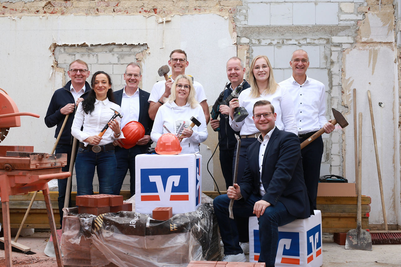 Mitarbeiter der Bank mit Baustellenhelmen und Werkzeugen auf einer Baustelle. 