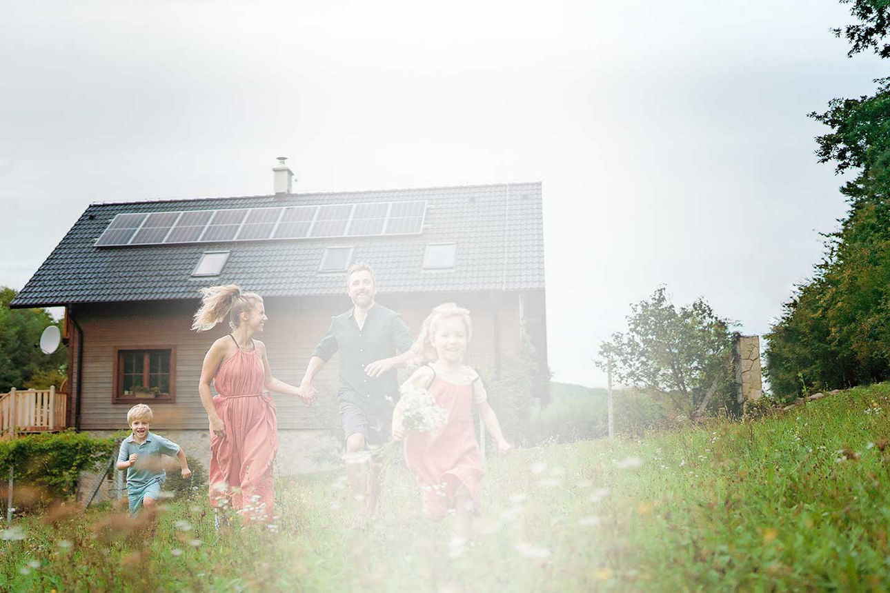Eine Familie mit zwei Erwachsenen und zwei Kindern läuft fröhlich über eine Wiese vor ihrem Haus.