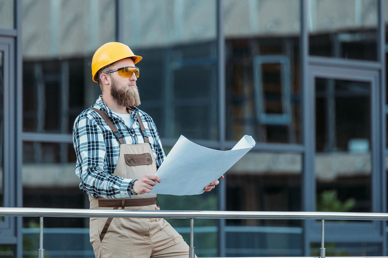 Ein Bauarbeiter mit gelben Helm und Sonnenbrille, der Baupläne auf einer Baustelle studiert.