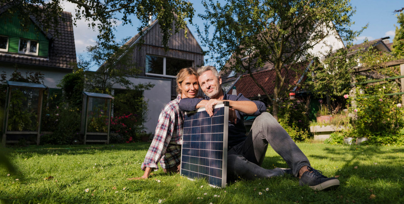 Mittelaltes Pärchen sitz auf eine Wiese vor einem Haus und hat ein Solarpanel in der Hand.