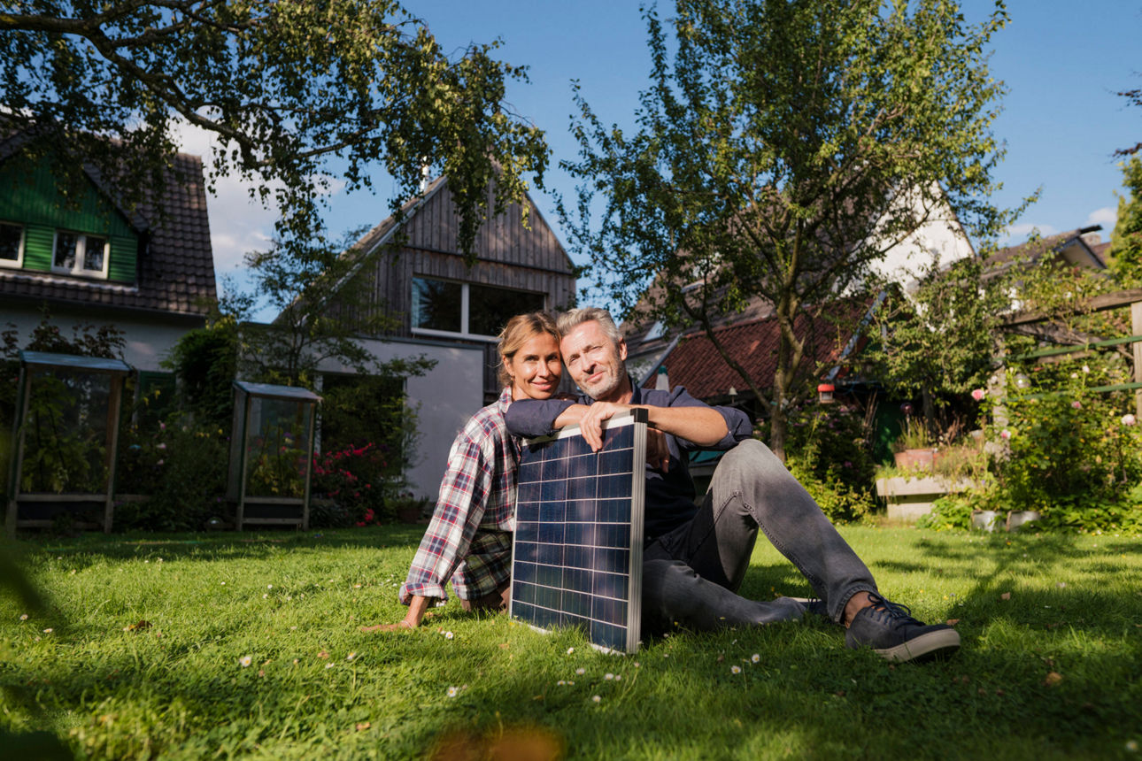 Pärchen mit Solarpanel in der Hand sitzt im Garten vor einem Haus
