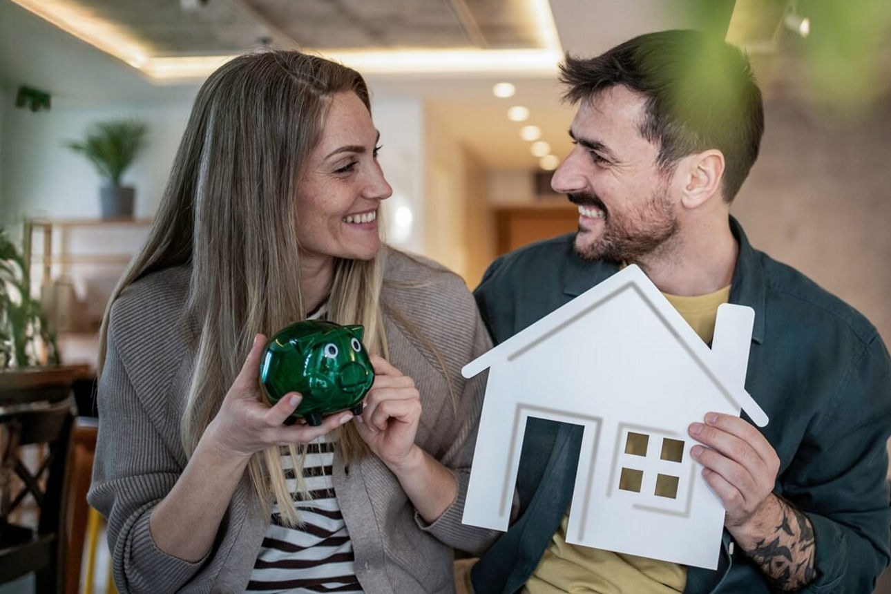 Paar stitzt im Wohnzimmer und hält ein grünes Sparschwein sowie ein weißes Hausmodell.