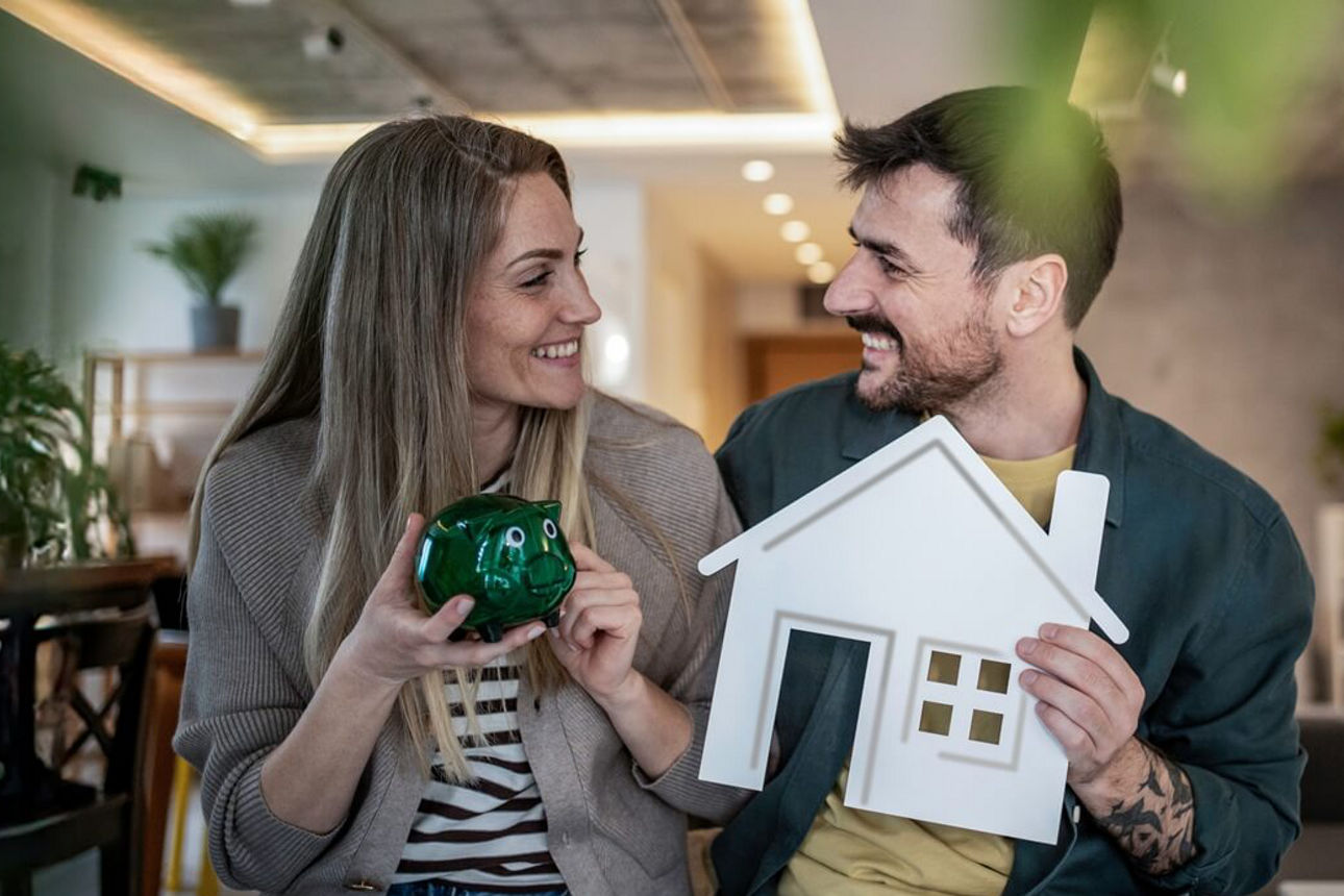 Paar stitzt im Wohnzimmer und hält ein grünes Sparschwein sowie ein weißes Hausmodell.