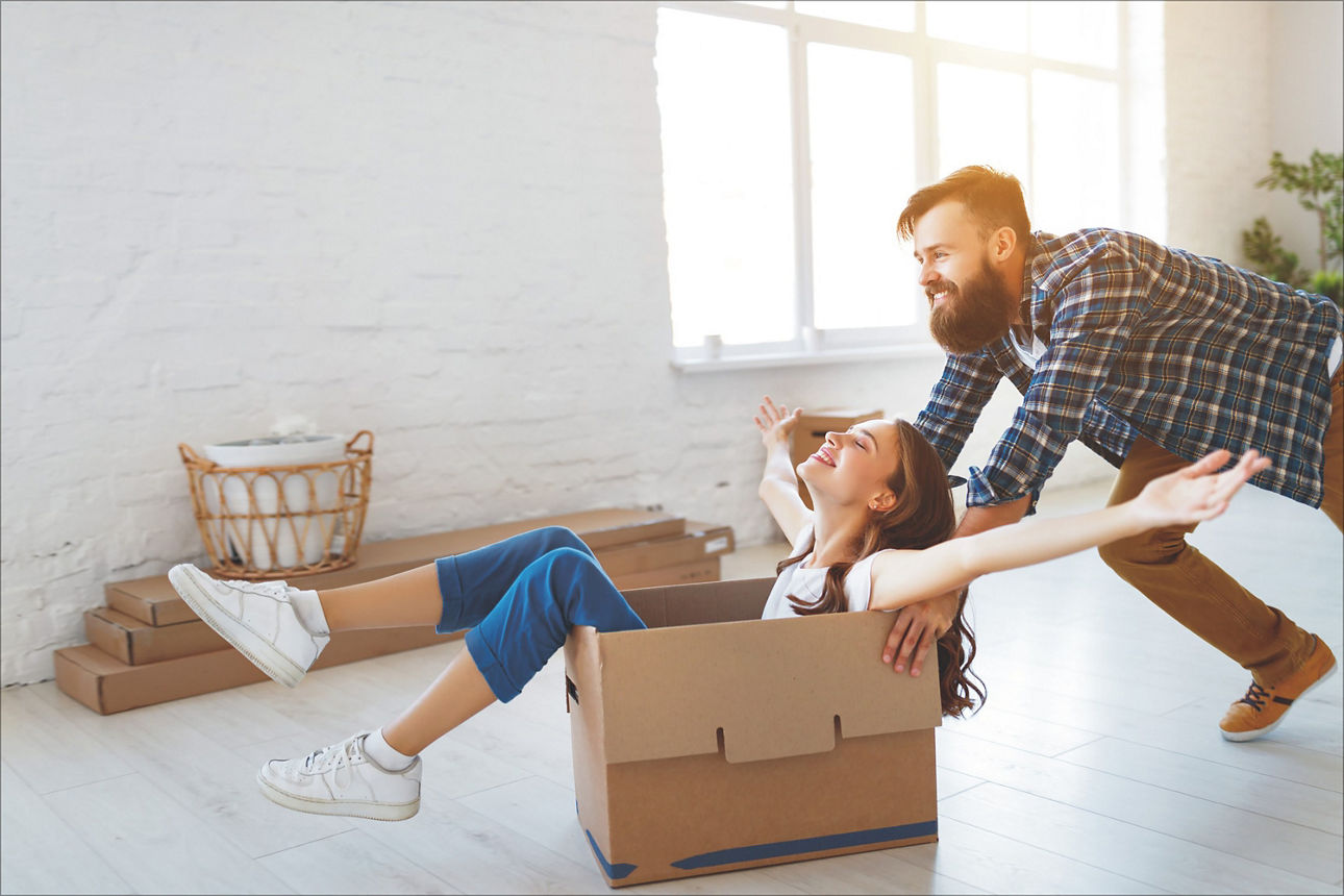 Mann der eine glückliche Frau in einem Karton durch eine wenig eingerichtete Wohnung schiebt