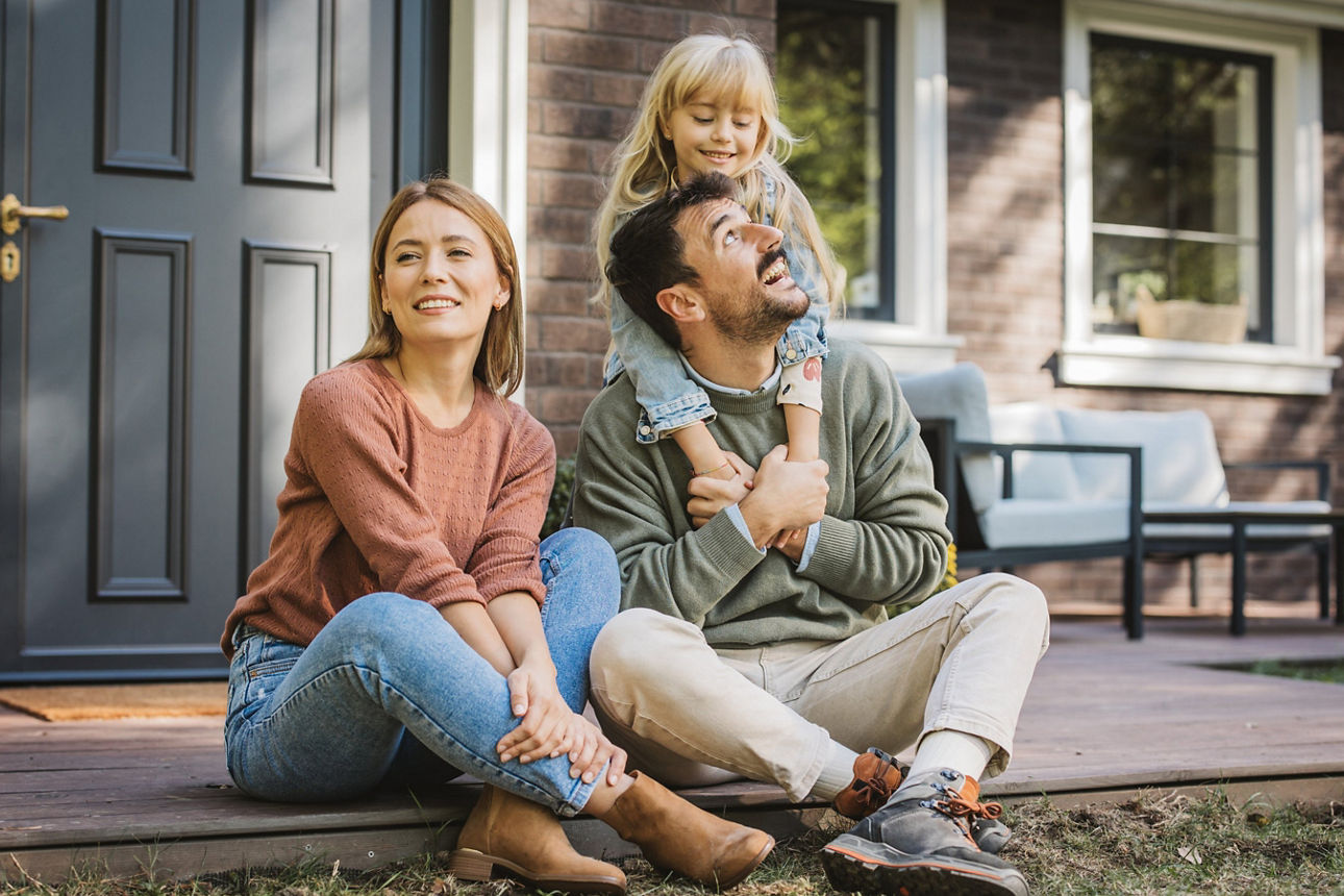 Junges Paar sitzt mit Kind vorm Eingang seines Eigenheims