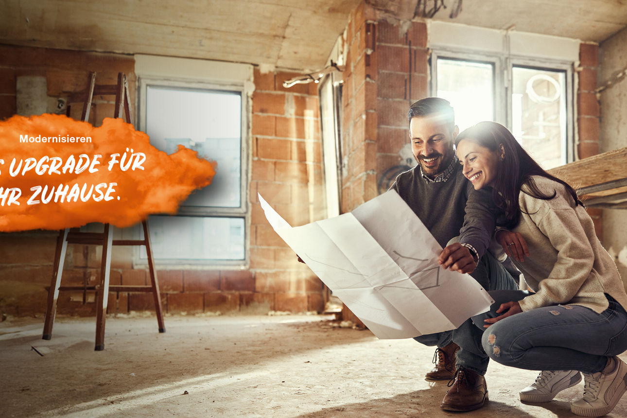 Junges Paar befinden sich in einer hockenden Position im Rohbau einer Wohnung und halten einen Bauplan in den Händen. Links im Bild steht auf einer orangenen Fläche "Modernisieren: Das Upgrade für Ihr Zuhause."