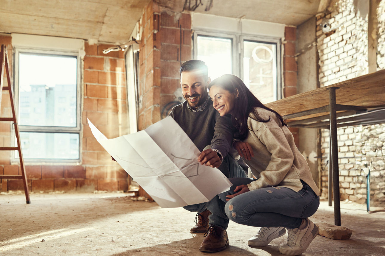 Junges Paar befinden sich in einer hockenden Position im Rohbau einer Wohnung und halten einen Bauplan in den Händen