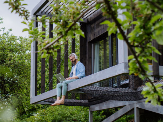 Mann sitzt mit Laptop und Kopfhörern auf einem Baumhaus