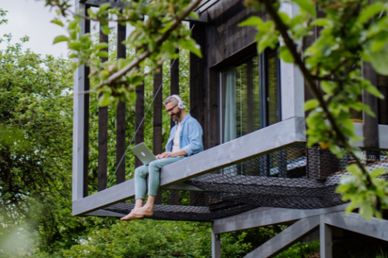 Mann sitzt mit Kopfhörern und Laptop auf dem Schoß in einem Baumhaus.
