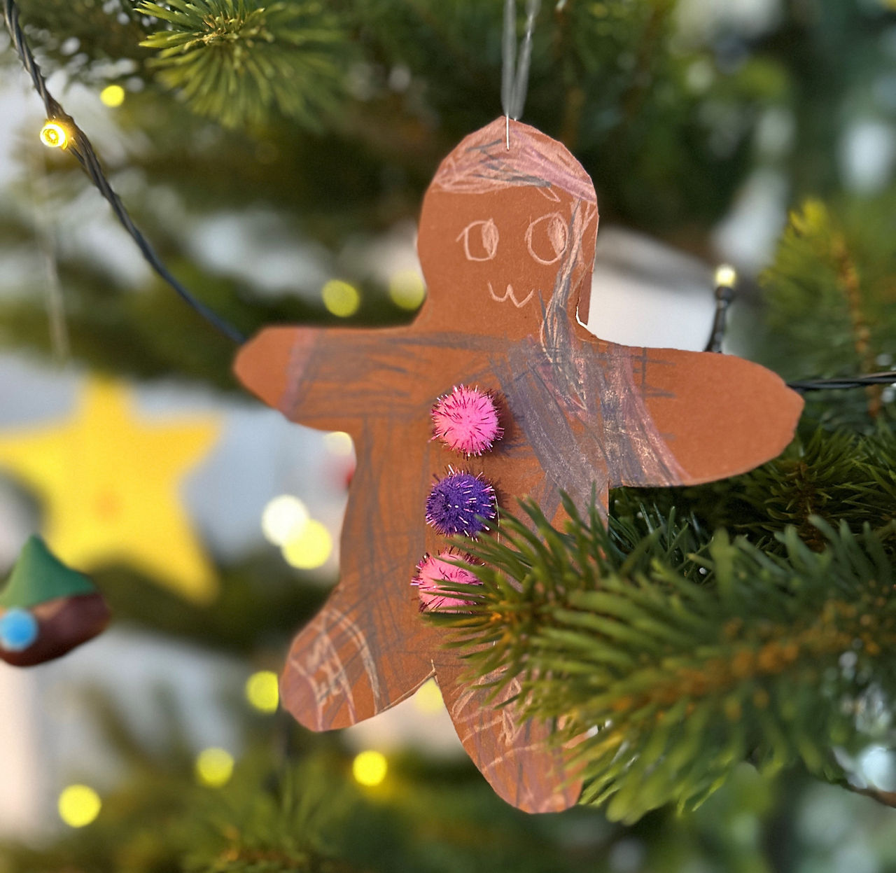 Braune Lebkuchenmann-Figur aus Papier mit drei bunten Pompons als Knöpfe hängt an einem Tannenzweig, im Hintergrund Lichterkette und unscharfe Weihnachtsdeko.