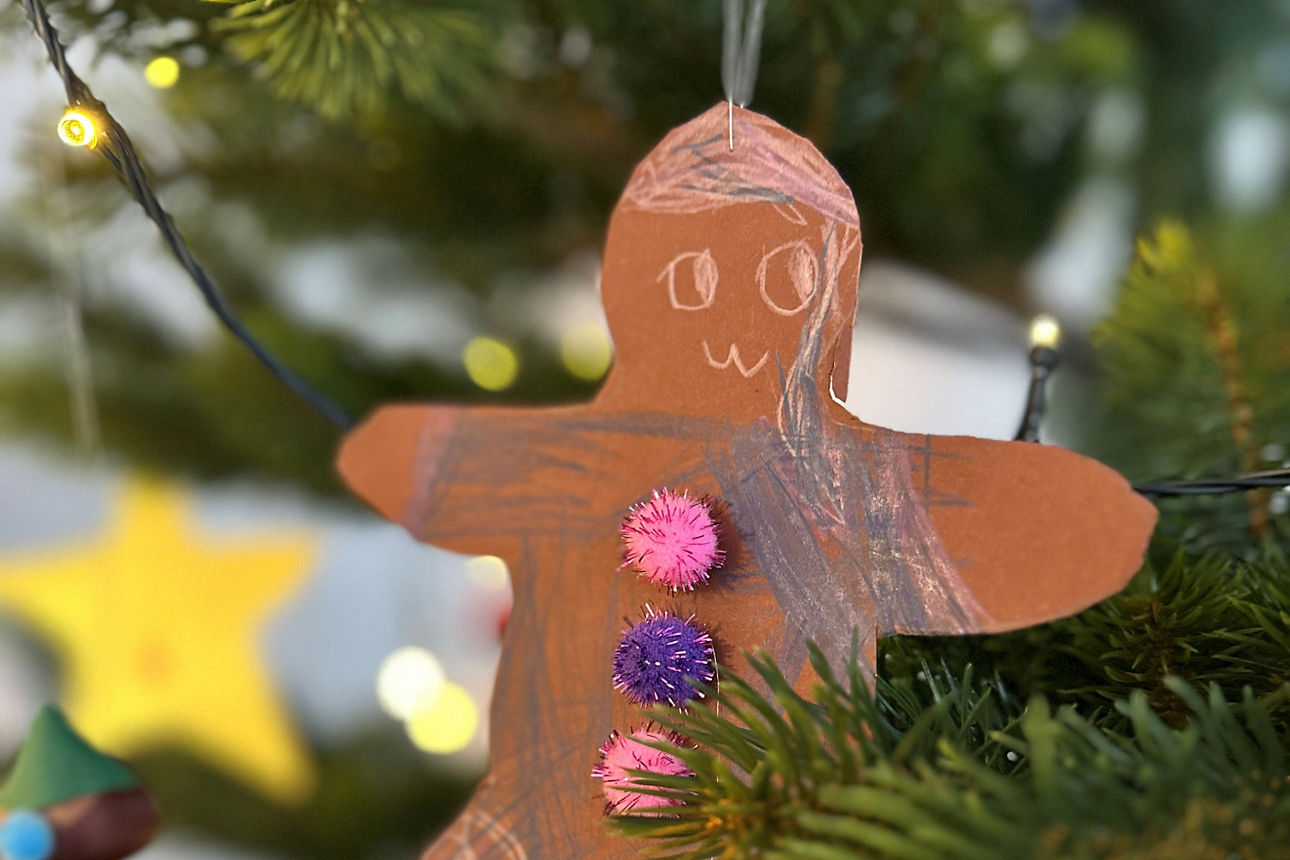 Braune Lebkuchenmann-Figur aus Papier mit drei bunten Pompons als Knöpfe hängt an einem Tannenzweig, im Hintergrund Lichterkette und unscharfe Weihnachtsdeko.