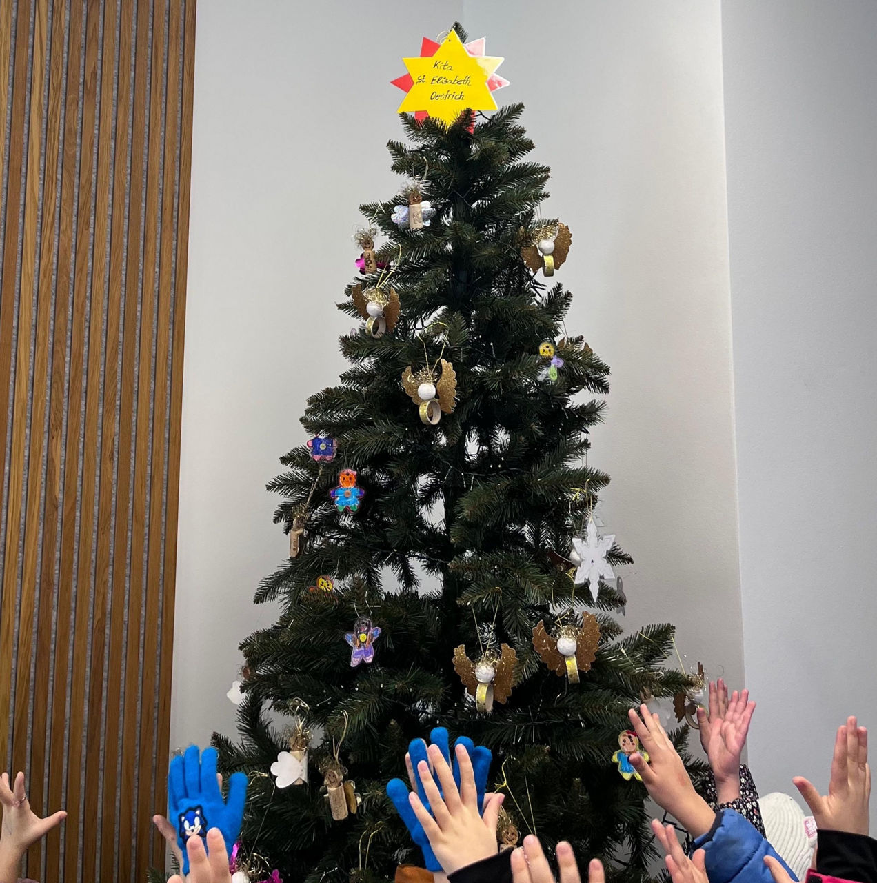 Weihnachtsbaum mit handgemachten Anhängern und gelbem Stern mit rotem Rand oben, davor mehrere erhobene Kinderhände.