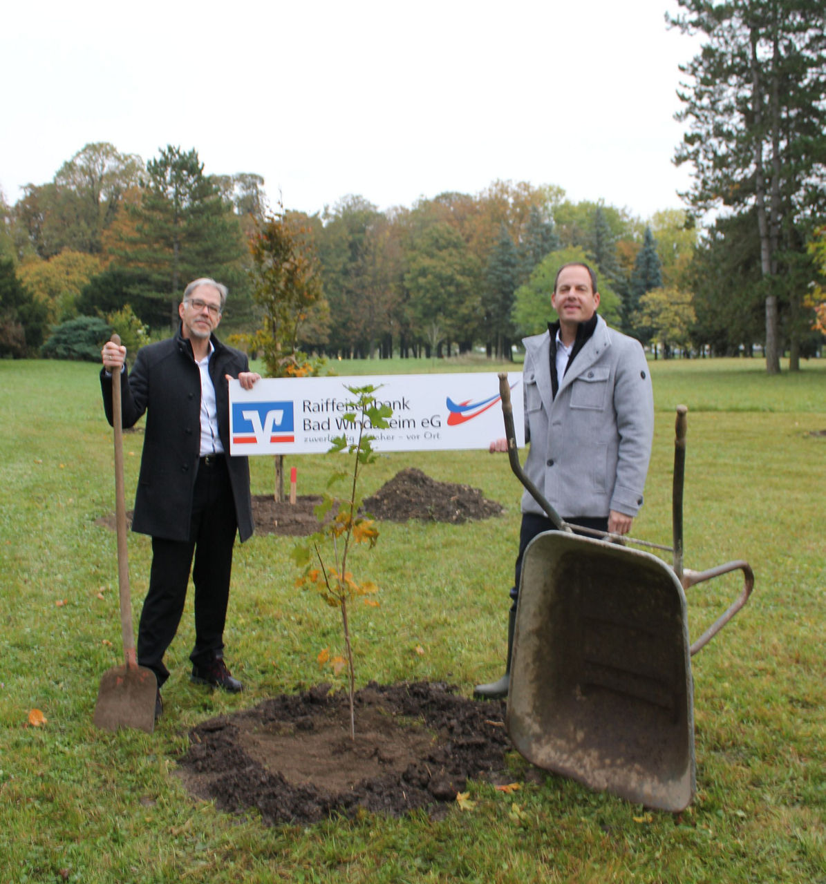Zwei Männer stehen links und rechts neben einem frich gepflanztem Baum halten ein Schild mit Logo der Bank in die Höhe