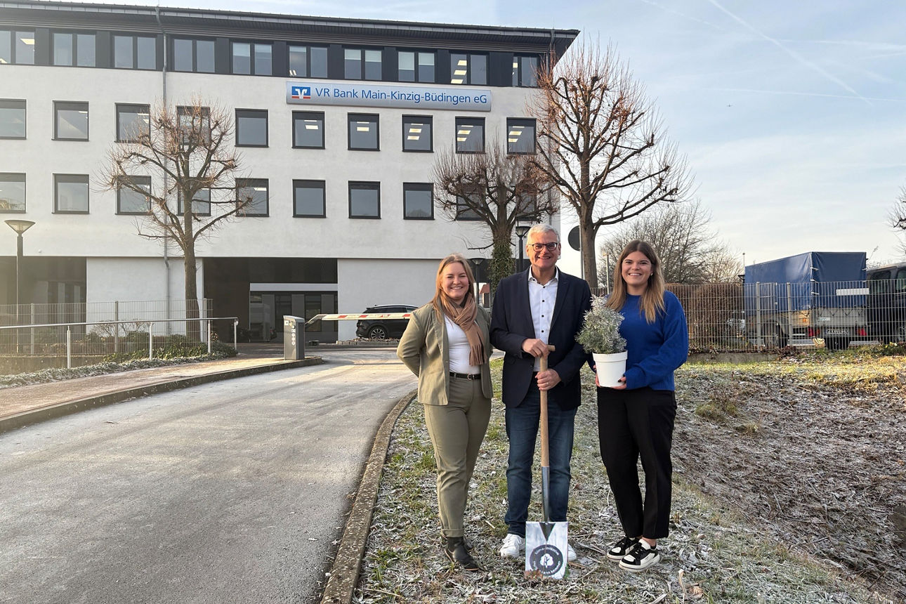Das Bild zeigt drei Personen vor dem VR Bank Main-Kinzig-Büdingen Gebäude in Linsengericht. Die Person in der Mitte hält einen Spaten, während die Frau auf der rechten Seite einen Baumsetzling hält. 
