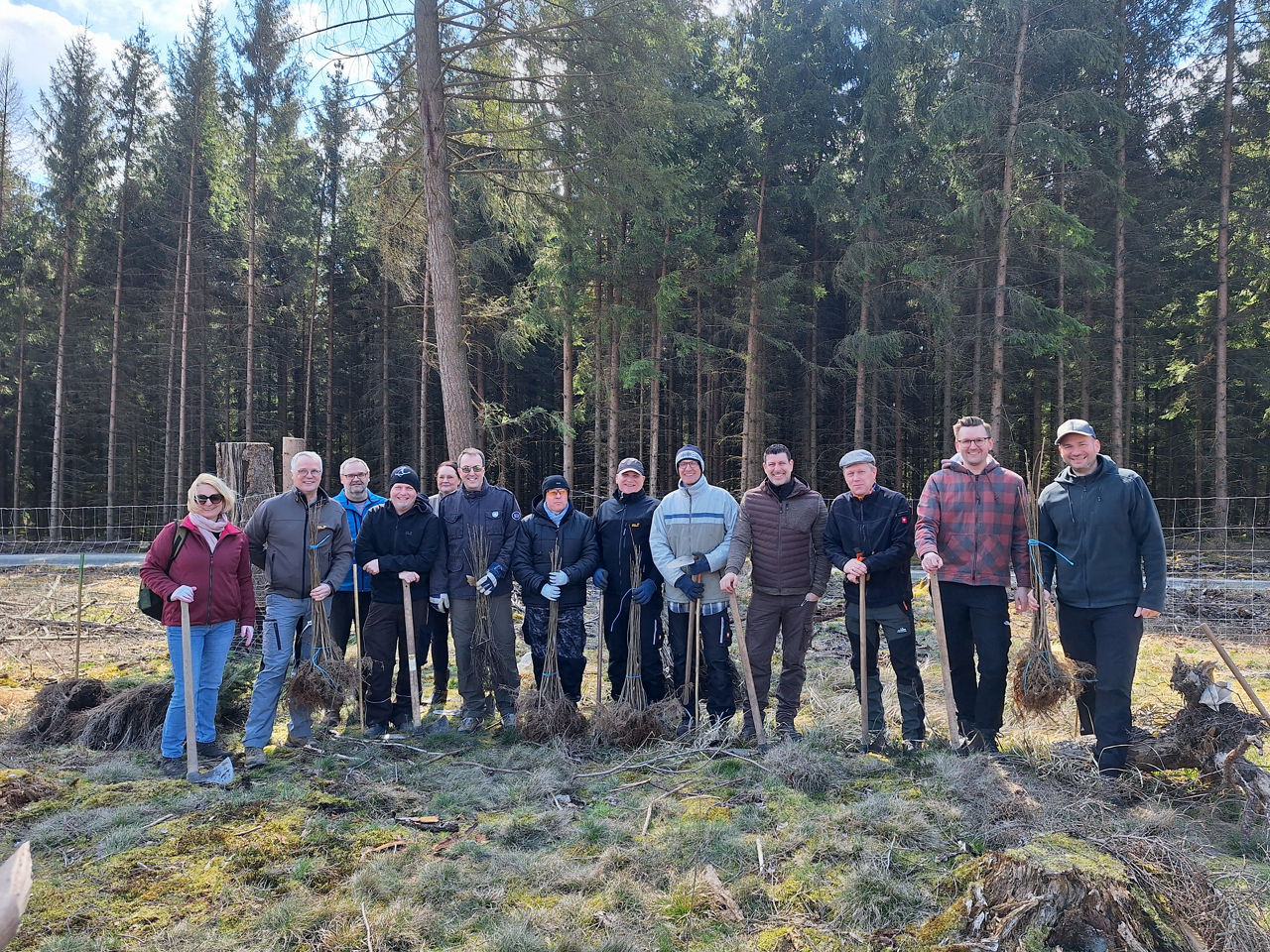 Auf dem Bild steht eine Gruppe von Menschen in einem Waldgebiet und hat Baumsetzlinge und Spaten in der Hand.