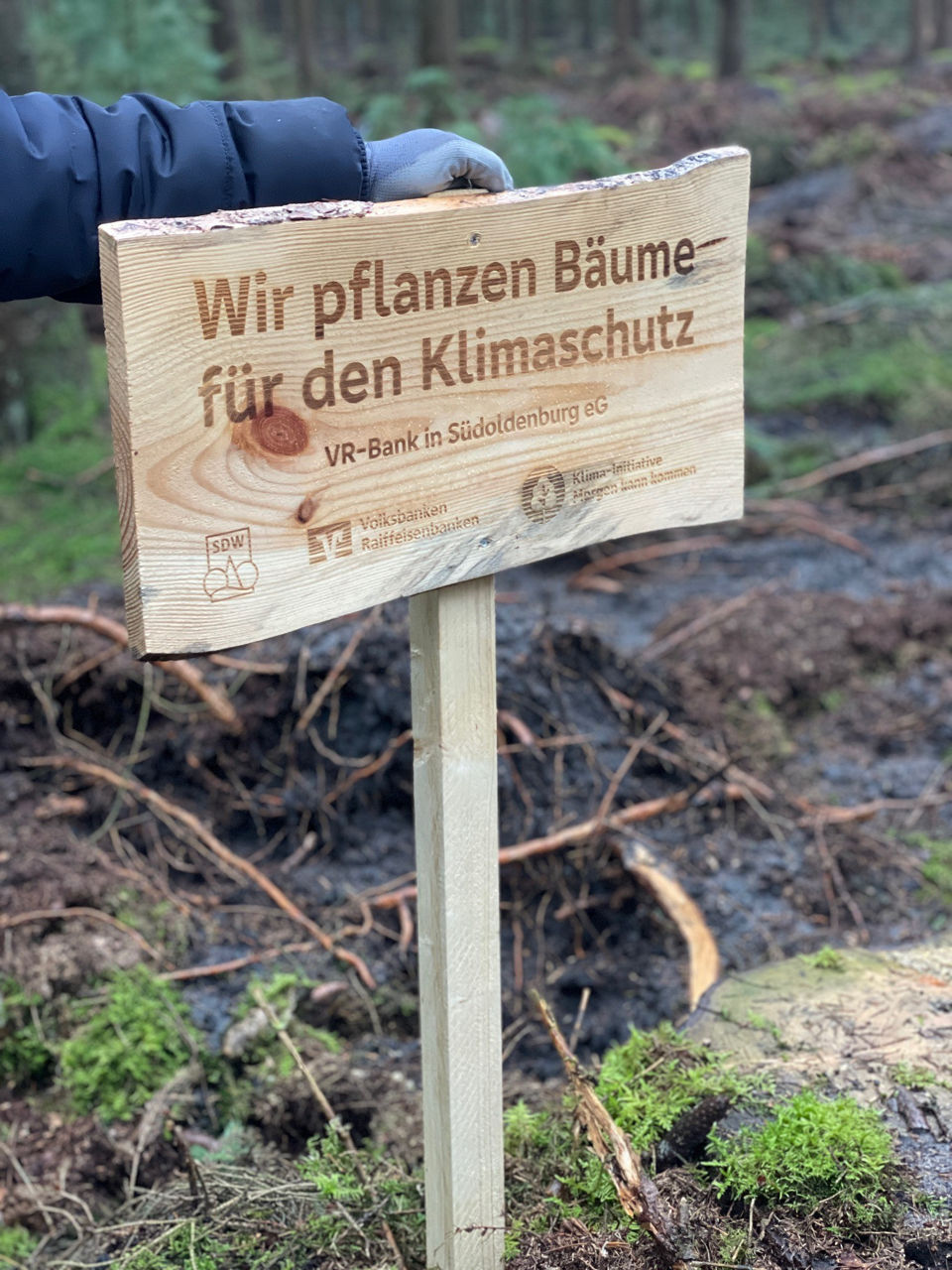 Mitarbeiter hält das Holzschild fest auf dem steht "Wir pflanzen Bäume für den Klimaschutz"