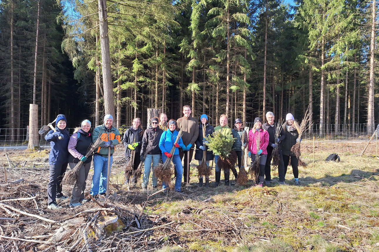 Auf dem Bild steht eine Gruppe von Menschen in einem Waldgebiet und hat Baumsetzlinge und Spaten in der Hand.