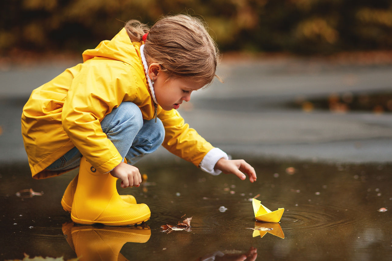 Ein Kind in gelbem Regenmantel und gelben Gummistiefeln, das sich über eine Pfütze beugt