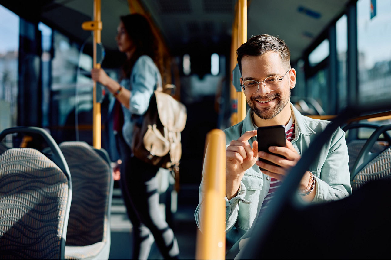 lächelnder junger Mann sitzt in Bus und schaut auf Handy