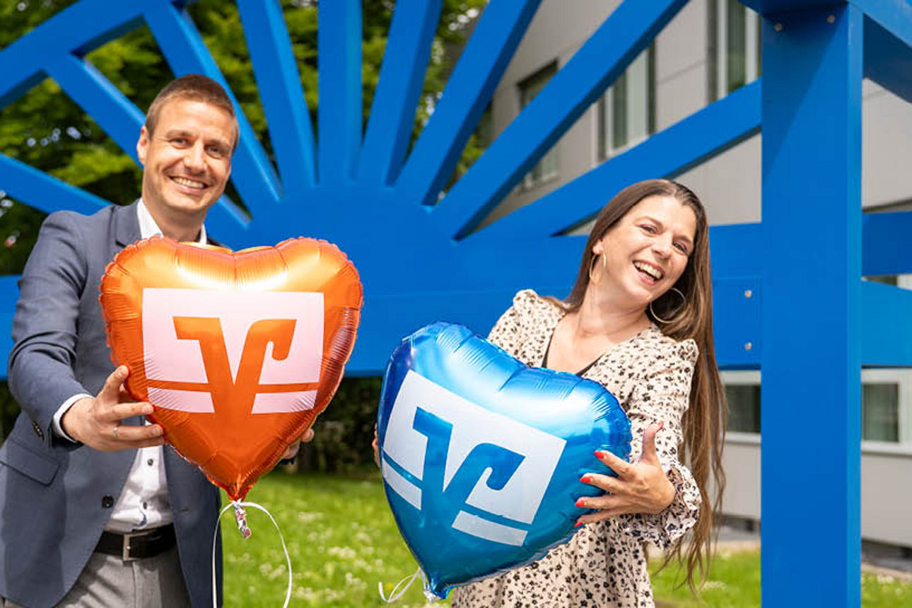 Ein Mann und eine Frau halte lächelnd eine Herzluftballon vor einer großen blauen Bank in der Hand