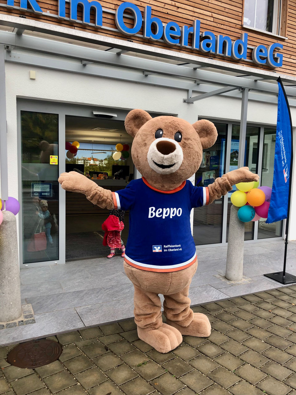 Das Bären-Bank-Maskottchen Beppo der Raiffeisenbank im Oberland steht, eingerahmt von bunten Luftballons, vor dem Eingang einer  Bankfiliale der Raiffeisenbank im Oberland