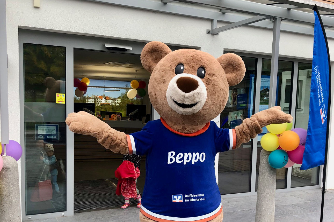 Das Bären-Bank-Maskottchen Beppo der Raiffeisenbank im Oberland steht, eingerahmt von bunten Luftballons, vor dem Eingang einer  Bankfiliale der Raiffeisenbank im Oberland