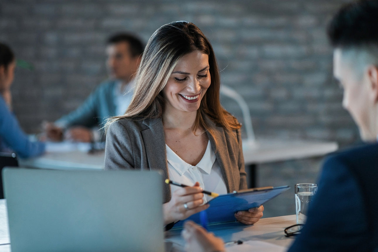 Zwei Personen sitzen an einem Tisch in einem Büro und betrachten gemeinsam ein Tablet neben Unterlagen und Brille.