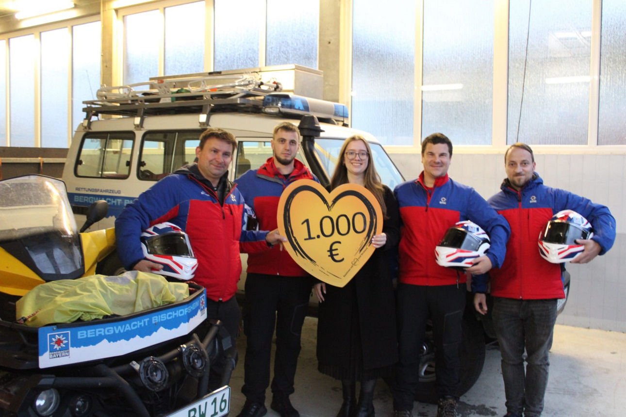 Auf dem Bild stehen vier Männer der Bergwacht Bischofsheim und in der Mitte eine Frau. Sie hält ein Herz in der Hand mit der Spendenaufschrift. Das Foto wurde vor einem Einsatzwagen der Bergwacht gemacht. 