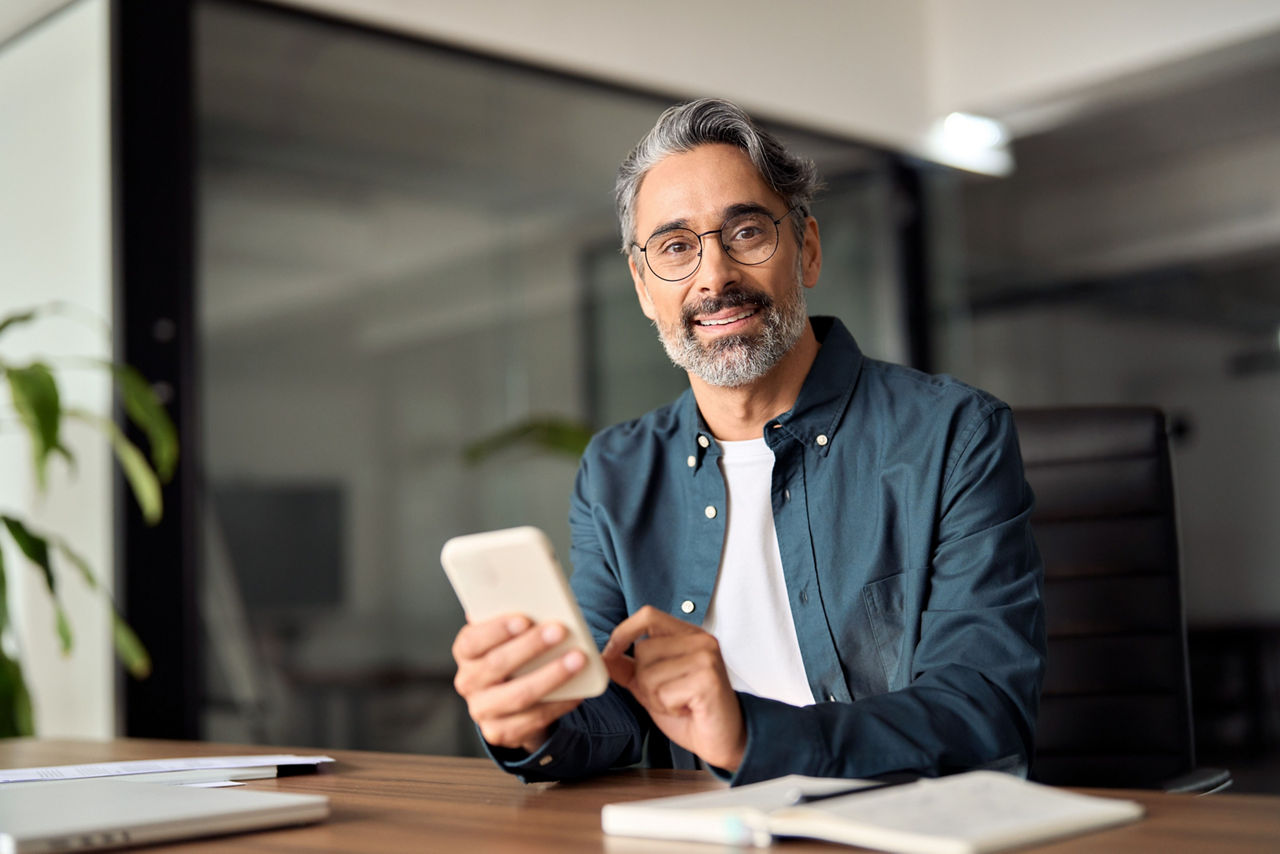 Mann im mitleren Alter am Schreibtisch lachdend  mit Handy in der Hand