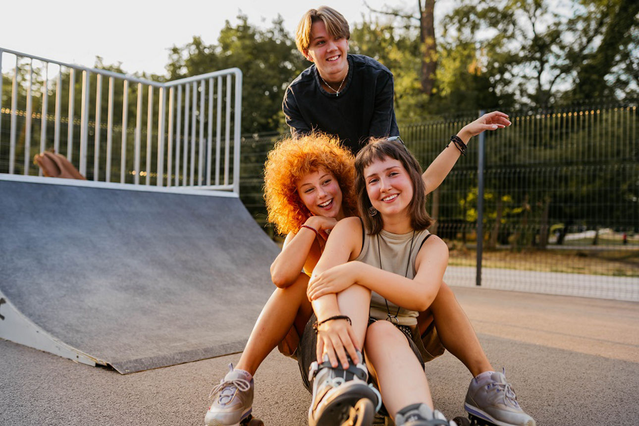 Drei junge Freunde sitzen auf dem Boden auf einer Skateanlage vor einer Rampe und lächeln in die Kamera