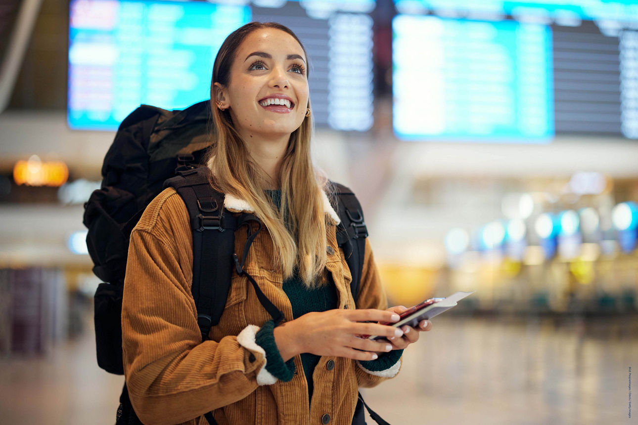 Mädchen steht lächeld mit Rucksack vor Anzeigentafel im Flughafen