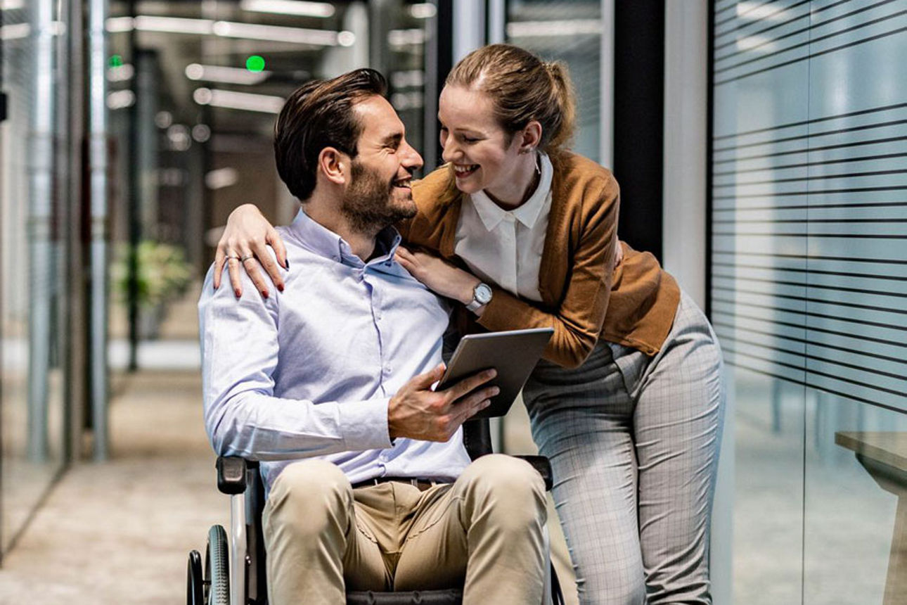 Frau unterstützt Mann im Rollstuhl im Büro, beide lächeln sich zu.