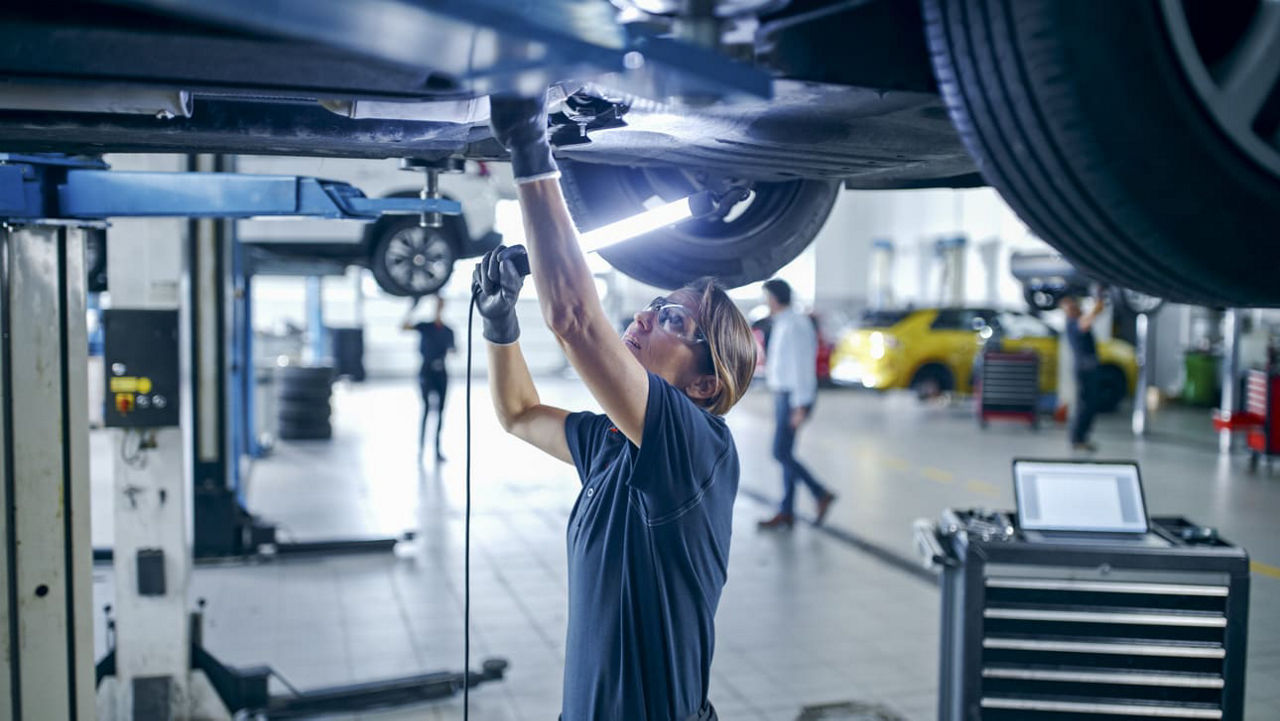 Eine junge Frau arbeitet in einer Kfz-Werkstatt unter einem Fahrzeug und inspiziert dieses