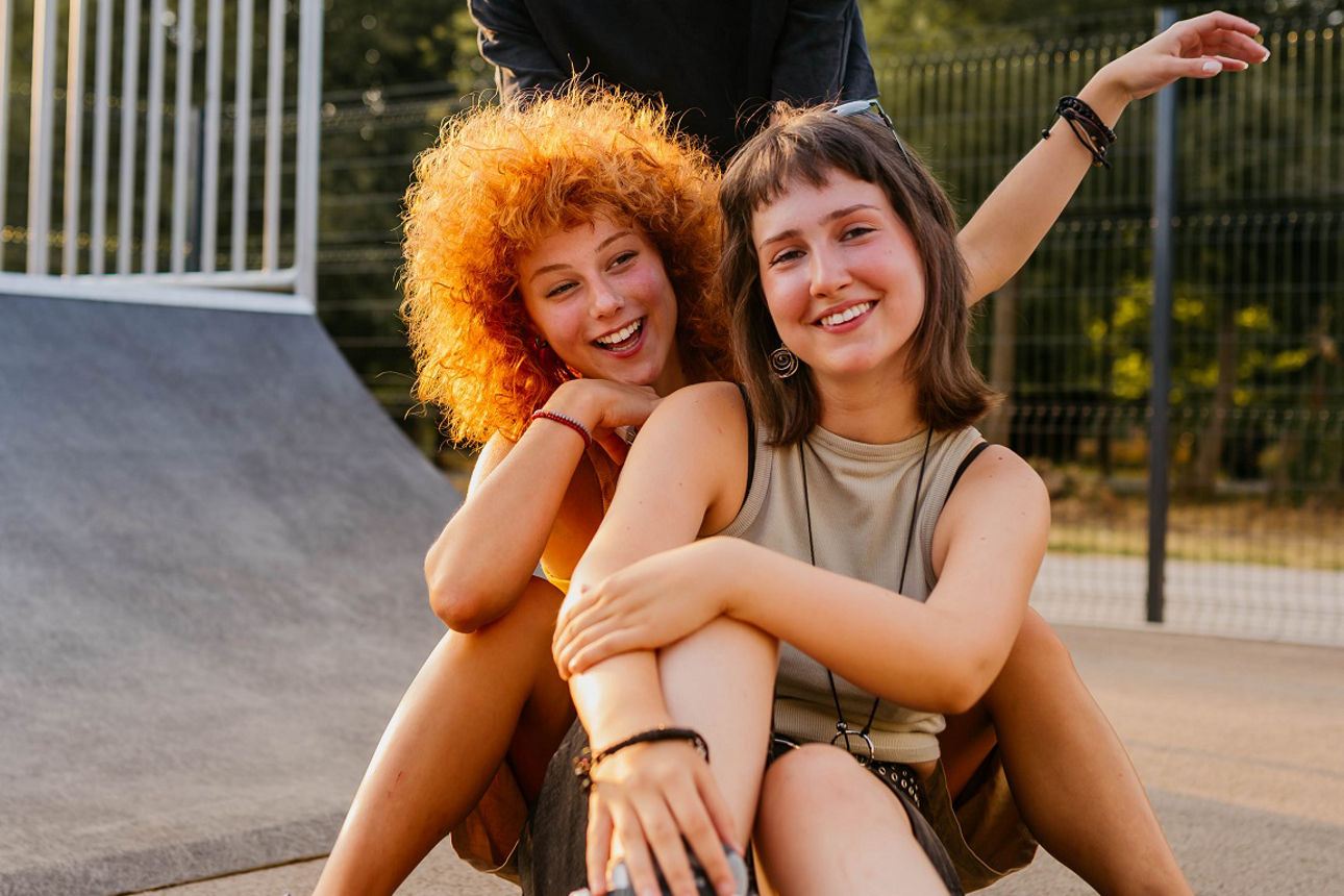 Drei junge Freunde sitzen auf dem Boden auf einer Skateanlage vor einer Rampe und lächeln in die Kamera