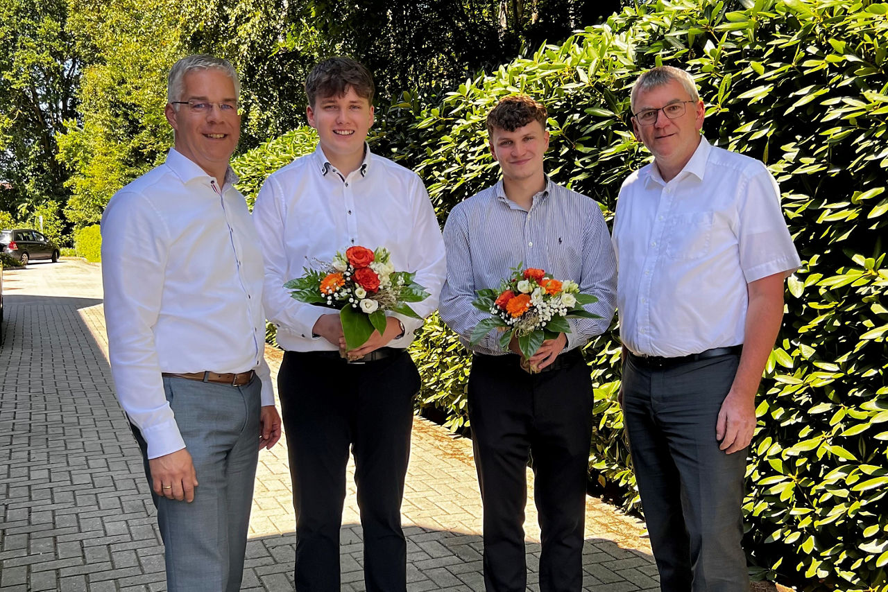 Vorstand Stefan Nowak, die beiden Prüflinge, mit Blumen und Ausbildungsleiter Mathias Lukassen draußen vor einer Hecke.