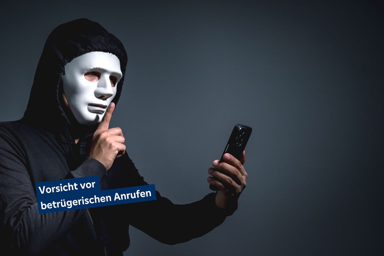 Ein maskierter Mann hält ein Telefon in der Hand und gleichzeitig einen Finger vor den Mund.