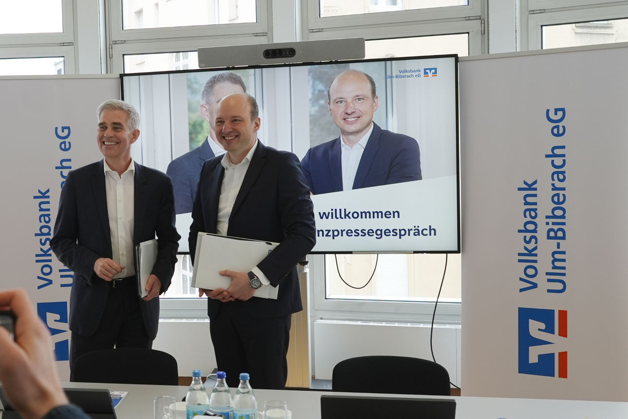 Beide Vorstände stehen vor einem Fernseher auf dem die Präsentation zu sehen ist.