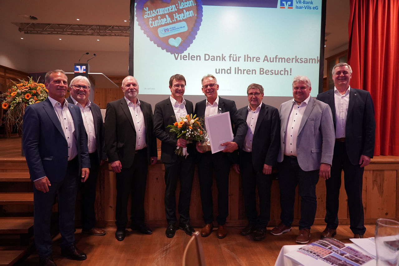 Acht Männer in Anzügen lächeln in die Kamera. Ein Mann hält einen Blumenstrauß und der Mann neben ihm eine Urkunde