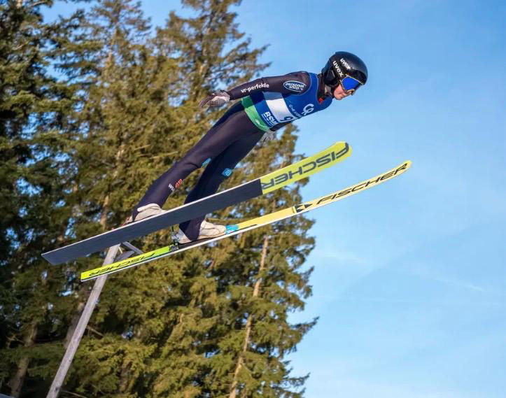 Skispringer in voller Ausrüstung fliegt dynamisch über die Schanze vor blauem Himmel und Waldkuliss
